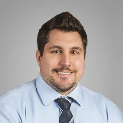 Staff member with short dark brown hair and a beard wearing a light blue shirt and dark patterned tie